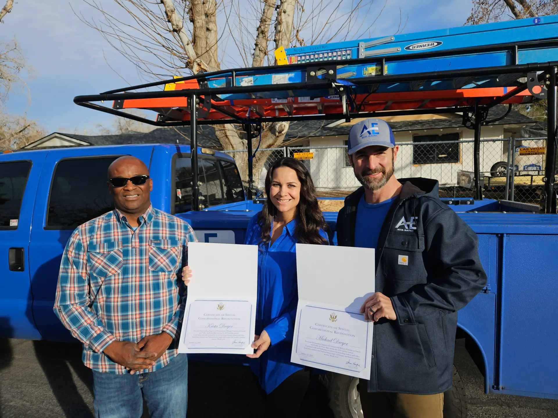 Three individuals, two holding certificates, standing in front of a blue truck with a rack.