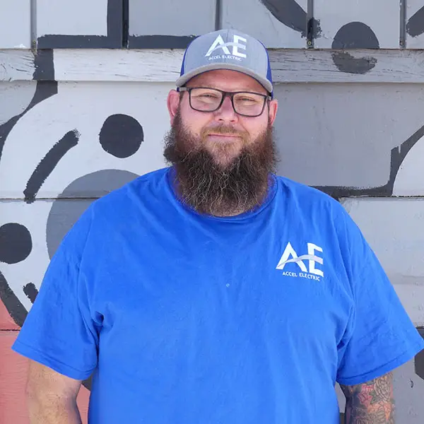 Man with a beard wearing a blue shirt and cap with the Accel Electric logo, standing against a painted wall.