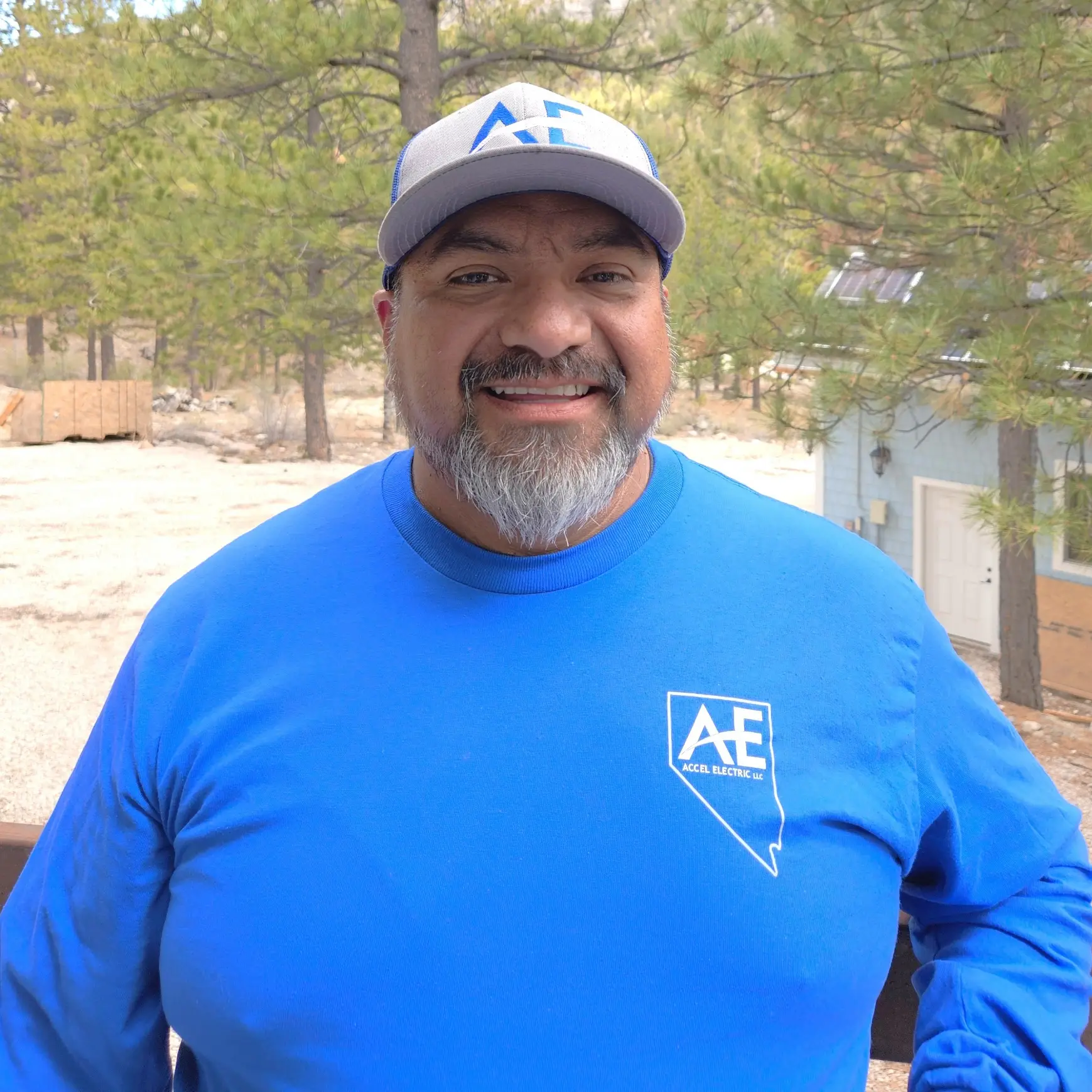 Man wearing a blue long sleeve shirt with 'AE' logo and a matching cap, standing outdoors with trees in the background.